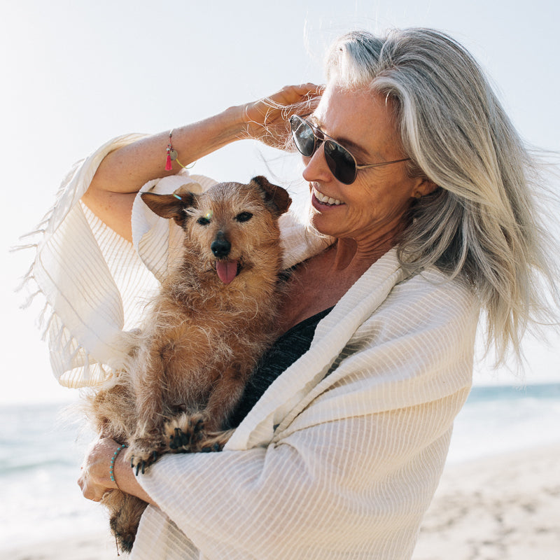 Lisa and her dog are enjoying a day at the beach. Lisa is wearing a Shoo for Good insect-repellant scarf to keep the bugs away. She's wearing the Spruce style in creamy cloud. Her dog is adorable.