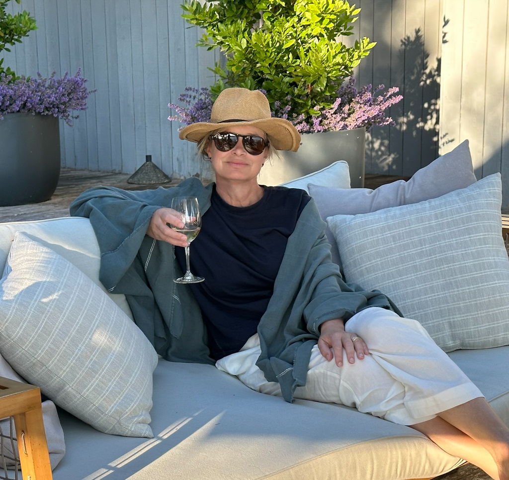 The slate green-gray cotton insect-repellent wrap repels ticks and mosquitoes. The woman wearing the insect-repellent wrap pairs it with a straw hat, sunglasses, and SFG tick-repellent cotton pants. She lounges on an outdoor daybed with a glass of wine in her hand.