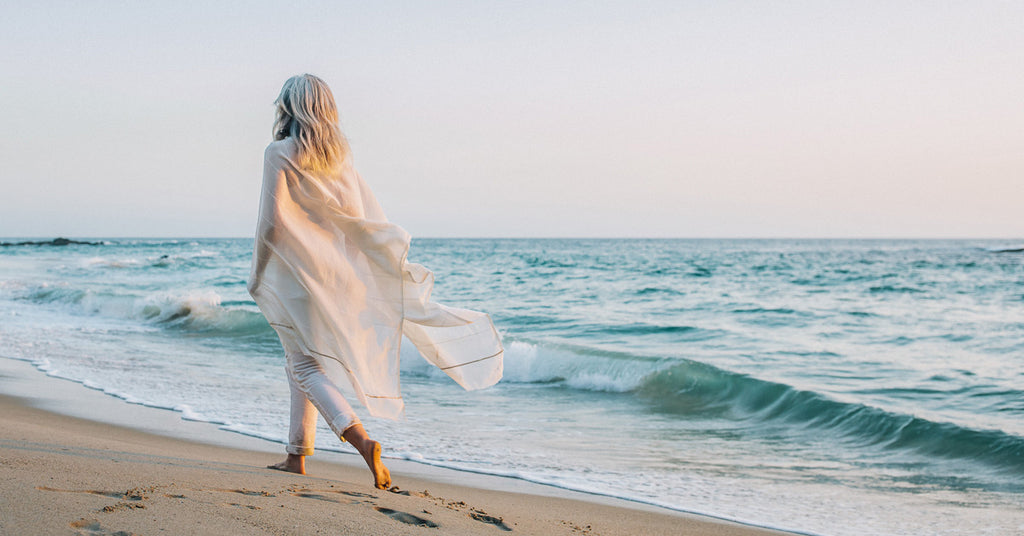 A woman is walking along the ocean wearing the Shoo For Good white Camellia wrap, and it blows in the breeze behind her. She looks very relaxed. We hope you try one for yourself. Love Shoo