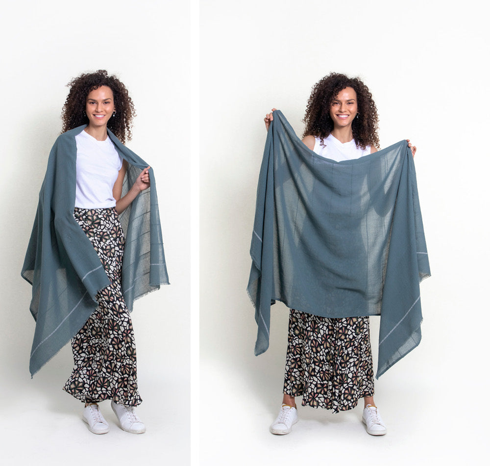 Shoo for Good's tick-repellent and mosquito-repellent oversize wrap in slate green-gray is held up by a model in a studio to show the dimensions and its sheer cotton. She pairs it with a white tank top, a long print skirt, and tennis shoes.