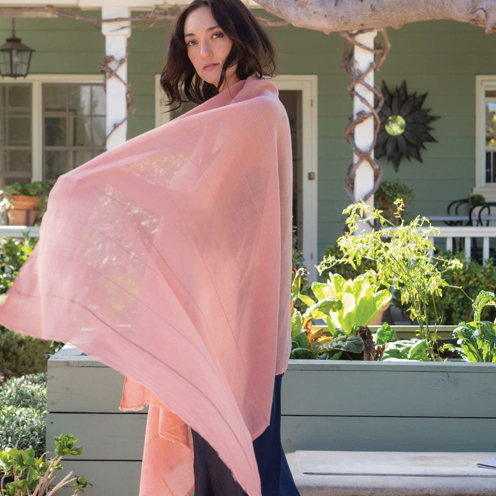 The mosquito-repellent Camellia wrap in blush with fine white stripes is lightweight, super soft, and keeps mosquitoes away. Permethrin is bound to the cotton fibers. The insect-repellency lasts for 70 washings, deterring insects for seasons. The lady wearing the wrap is relaxed in the garden, knowing bugs won't bother her. Insect Shield technology keeps bugs away.